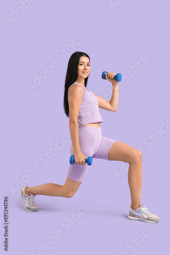 Sporty young woman training with dumbbells on lilac background