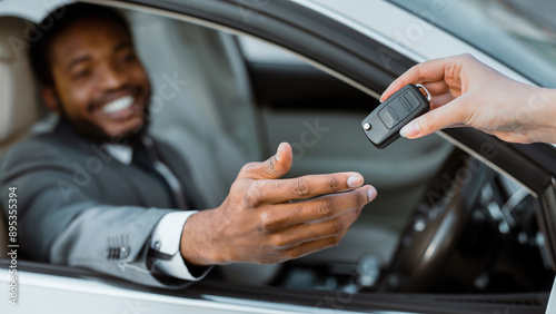 A man sits in the drivers seat of a new car, extending his hand to receive the keys from someone outside the vehicle. He is smiling and appears to be happy and excited about his new car. photo