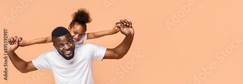A joyful black father and daughter are playing together in a studio setting. The father is holding the daughter on his back, and they are both smiling and laughing, panorama with copy space