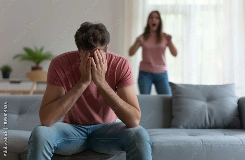 Young man sitting couch in front, crying and covering his face with hands while woman standing behind him is shouting at her husband. Domestic abuse concept Young man sitting couch in front, crying and covering his face with hands while woman standing behind him is shouting at her husband. Domestic abuse concept