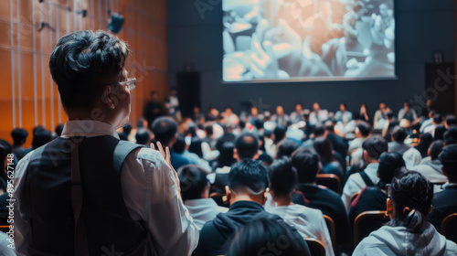 A large audience attentively listens to a speaker in a dimly lit conference hall, reflecting a moment of collective focus and engagement. © VK Studio