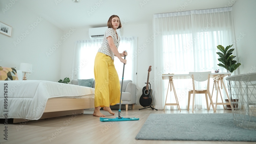Caucasian housekeeper mopping the floor while dancing in lively mood at modern apartment. Happy energetic mother or cleaner enjoy cleaning the living room while moving to music. Lifestyle. Pedagogy. Caucasian housekeeper mopping the floor while dancing in lively mood at modern apartment. Happy energetic mother or cleaner enjoy cleaning the living room while moving to music. Lifestyle. Pedagogy.
