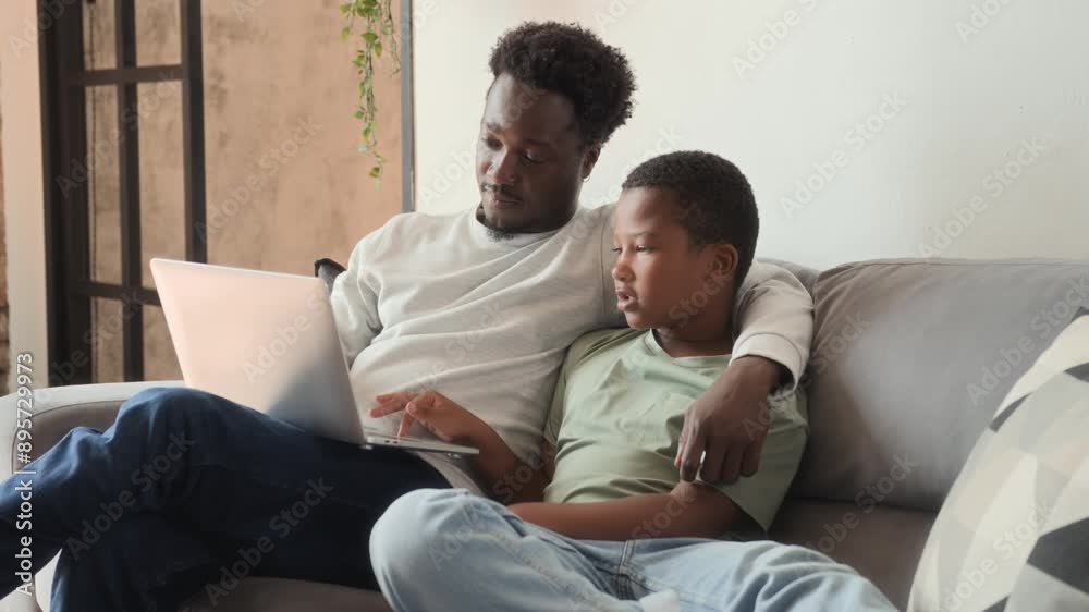 African American father and boy sitting together on couch and using laptop. Black skinned man hugs his son, typing on laptop keyboard researching online and browsing African American father and boy sitting together on couch and using laptop. Black skinned man hugs his son, typing on laptop keyboard researching online and browsing