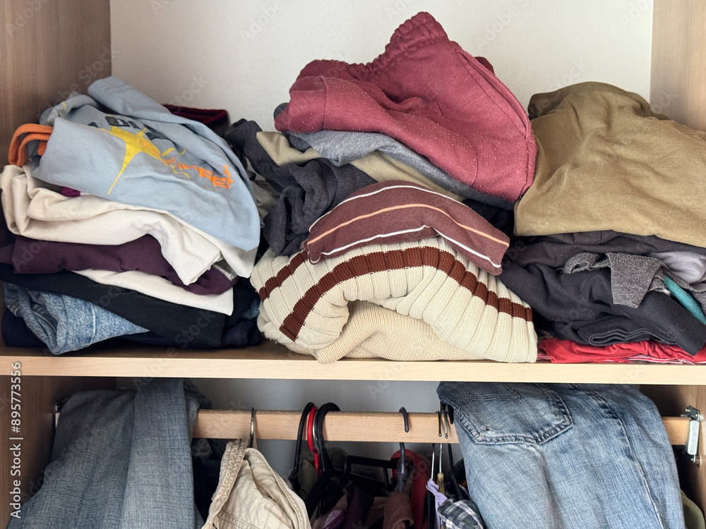 Messy wardrobe with assorted casual clothes, sweaters, and pants on shelves and hangers Messy wardrobe with assorted casual clothes, sweaters, and pants on shelves and hangers