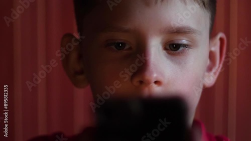 Caucasian boy 9-10 years old looks with horror and bewilderment at the phone screen in the dark, covering his mouth with his hand, obscene content