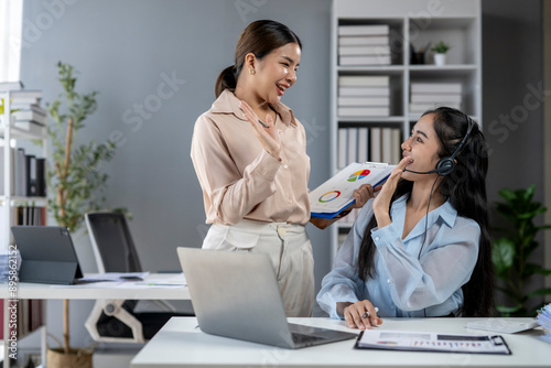 Two women are sitting at a desk with a laptop and a stack of papers