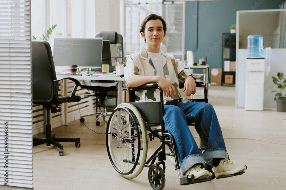 Portrait of modest highly experienced programmer with disability sitting for photo in center of modern office fully equipped with electronic devices and comfort workspaces Portrait of modest highly experienced programmer with disability sitting for photo in center of modern office fully equipped with electronic devices and comfort workspaces