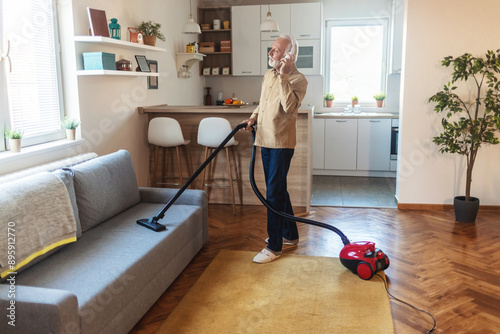 Mature man having fun vacuuming the carpet and sofa in the room, listening to music on wireless headphones and dancing with the vacuum cleaner.