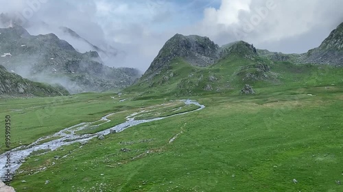 Une petite rivière serpente à travers une plaine de montagne, entourée de sommets majestueux. L'eau scintille sous le ciel clair, offrant un contraste paisible avec les montagnes environnantes.