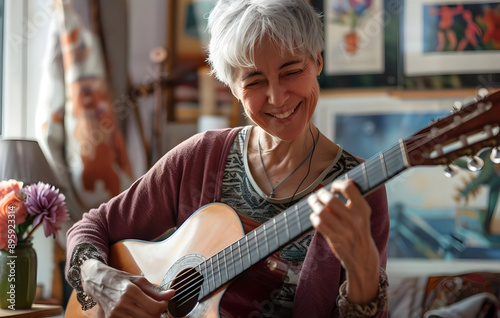 Smiling elderly woman playing an acoustic guitar in a cozy room filled with paintings and natural light, enjoying music. photo