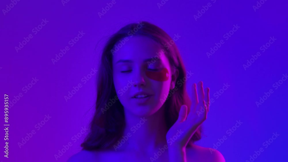 Young woman posing at camera puts an eyepatches under eyes, isolated on blue neon background. Natural beauty and skin care concept Young woman posing at camera puts an eyepatches under eyes, isolated on blue neon background. Natural beauty and skin care concept