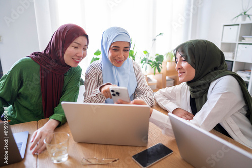 Three women in hijabs share a moment of laughter while looking at a smartphone in a modern office Their interaction highlights a blend of professionalism and friendship