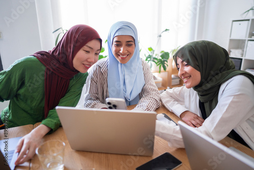 Three women in hijabs share a moment of laughter while looking at a smartphone in a modern office Their interaction highlights a blend of professionalism and friendship