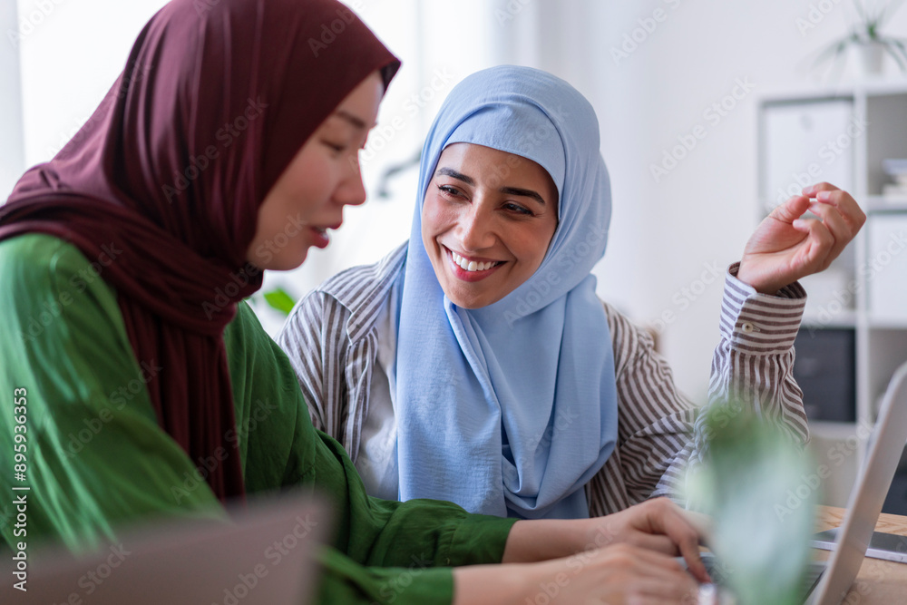 Two women in hijabs collaborate on a project in a modern office space One woman in a green hijab is smiling while the other in a blue hijab focuses on their work Two women in hijabs collaborate on a project in a modern office space One woman in a green hijab is smiling while the other in a blue hijab focuses on their work