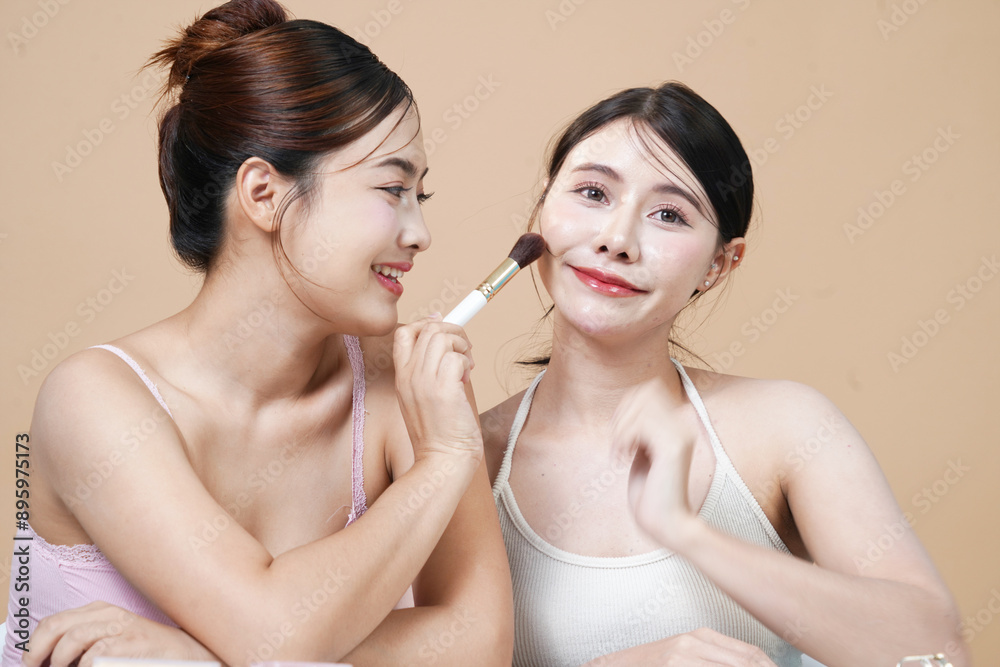 Two young Asian women, 23 years old, beautiful women, holding blush brushes, applying makeup together, bright facial skin, Korean style, matte cream background The concept of beautiful, healthy skin Two young Asian women, 23 years old, beautiful women, holding blush brushes, applying makeup together, bright facial skin, Korean style, matte cream background The concept of beautiful, healthy skin