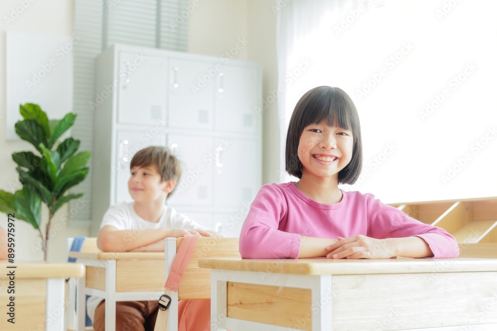 Smiling Asian schoolgirl sits at her desk in a classroom with other students, looking directly at the camera.