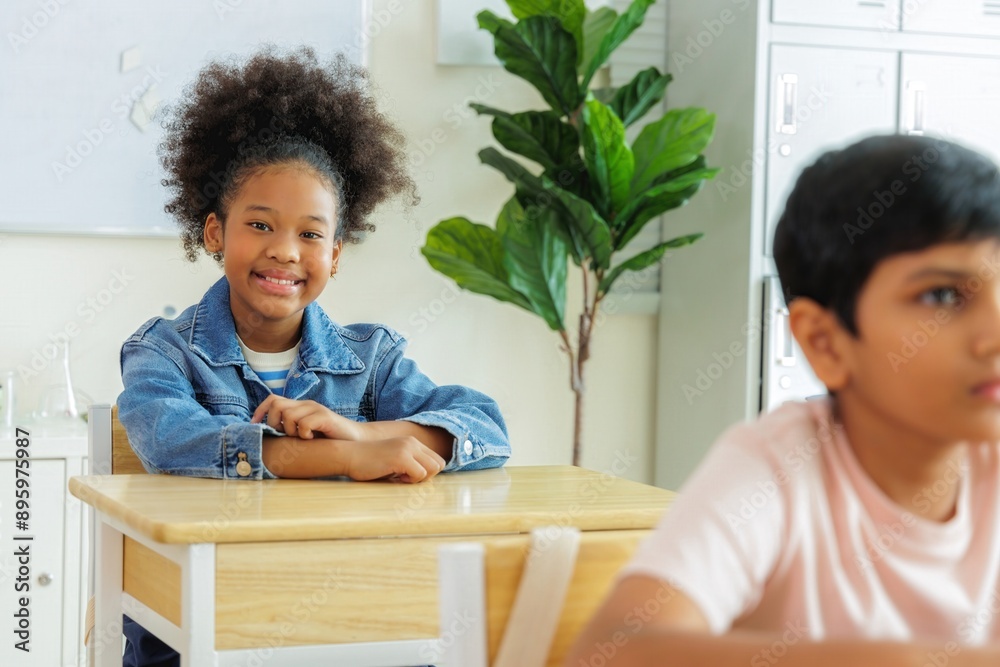 Fototapeta premium Smiling young girl in denim jacket sits at a desk in a classroom. Her classmate is sitting next to her.