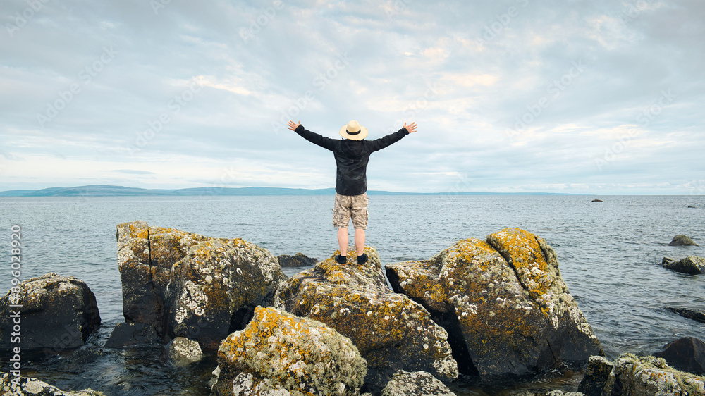 Handsome bearded man with straw hat standing on the rocks by the ocean at wild Atlantic way, adventure and lifestyle concept, summer, vacation, nature background