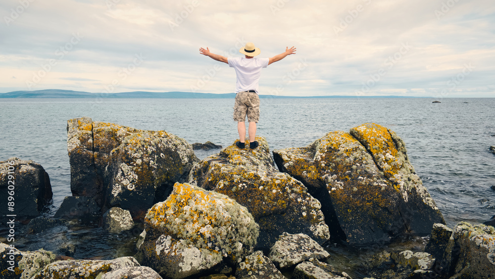 Handsome bearded man with straw hat standing on the rocks by the ocean at wild Atlantic way, adventure and lifestyle concept, summer, vacation, nature background