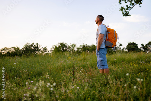 A male tourist with a backpack on his shoulders is standing in a meadow, looking ahead.