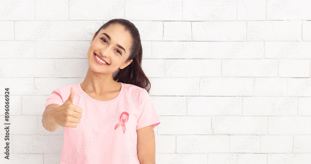 Breast Cancer Recovery. Woman In T-Shirt With Pink Ribbon Gesturing Thumbs Up Posing Over White Brick Wall Background. Selective Focus Breast Cancer Recovery. Woman In T-Shirt With Pink Ribbon Gesturing Thumbs Up Posing Over White Brick Wall Background. Selective Focus