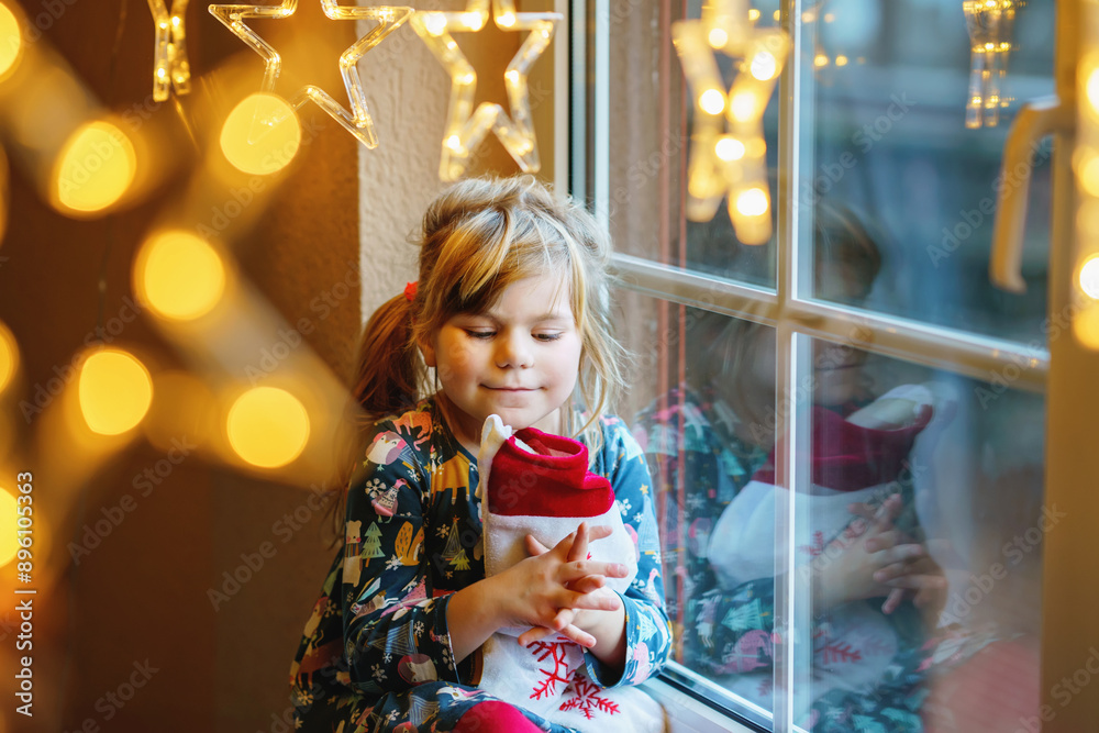 Little preschool girl holding cup Santa Claus boot with gift called Nikolausstiefel in German. Happy child wait on holiday by window with Christmas lights in winter. Cozy family celebration of xmas. Little preschool girl holding cup Santa Claus boot with gift called Nikolausstiefel in German. Happy child wait on holiday by window with Christmas lights in winter. Cozy family celebration of xmas.