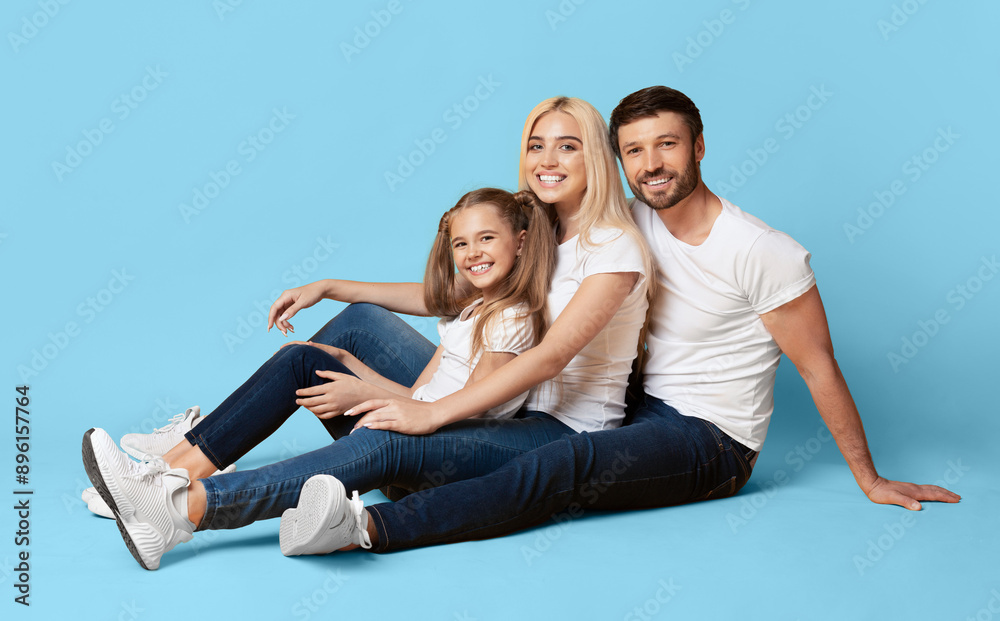 A family of three sits on a light blue background, smiling and looking at the camera. They are all wearing casual clothing A family of three sits on a light blue background, smiling and looking at the camera. They are all wearing casual clothing
