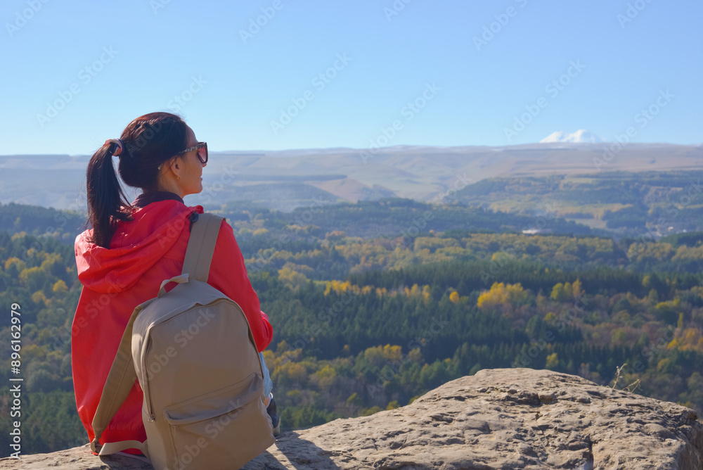 A girl tourist with a backpack sits on the top of a mountain and enjoys the beautiful views. A walking woman in an orange jacket rests on a rock, looking at the beautiful sunny landscape of the