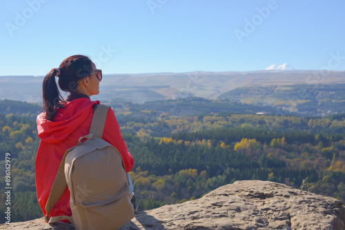 Obraz na płótnie A girl tourist with a backpack sits on the top of a mountain and enjoys the beautiful views. A walking woman in an orange jacket rests on a rock, looking at the beautiful sunny landscape of the w salonie