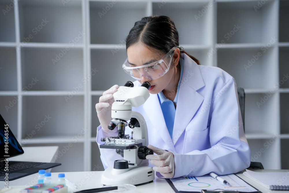 Young researcher in a lab coat and safety glasses conducting research with a microscope in a state of the art laboratory Young researcher in a lab coat and safety glasses conducting research with a microscope in a state of the art laboratory