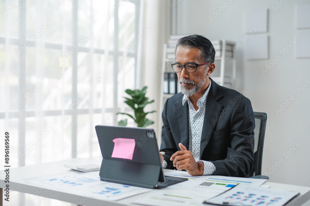 Mature businessman immersed in work at his office desk, focused on his laptop in a bright, modern workplace flooded with natural light Mature businessman immersed in work at his office desk, focused on his laptop in a bright, modern workplace flooded with natural light