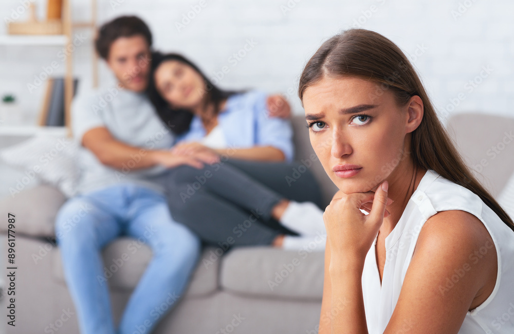 Jealousy. Unhappy Girl Looking At Camera While Her Dating Friends Hugging Sitting On Sofa Indoor. Selective Focus Jealousy. Unhappy Girl Looking At Camera While Her Dating Friends Hugging Sitting On Sofa Indoor. Selective Focus
