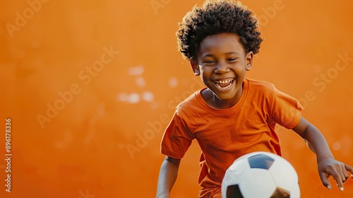 A delighted boy in an orange shirt is chasing after a soccer ball, laughing as he runs. The bright orange background highlights his energetic and fun-loving nature