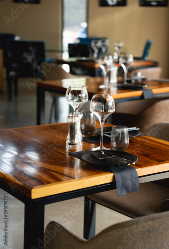 A close-up photo of a meticulously set table in a modern restaurant. The table features a rich, dark wooden surface with a metal frame. A black napkin and a black plate are placed on the table, with s