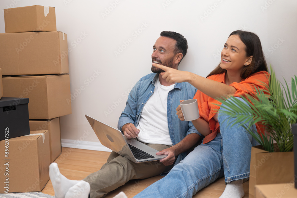 Young guy sits on floor with laptop with happy wife drinking tea from cup pointing finger at various cardboard boxes for arrangement of modern house Young guy sits on floor with laptop with happy wife drinking tea from cup pointing finger at various cardboard boxes for arrangement of modern house