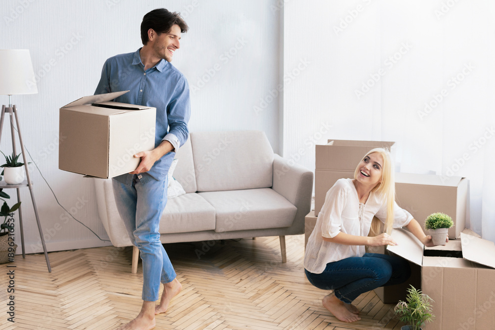 A young man carries a box in his arms and smiles at a young woman sitting on the floor unpacking boxes in a new apartment. A young man carries a box in his arms and smiles at a young woman sitting on the floor unpacking boxes in a new apartment.