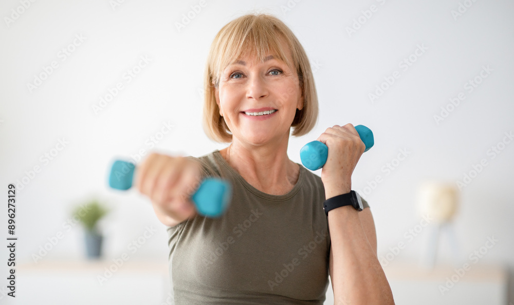 Home training concept. Strong senior woman doing exercises with dumbbells indoors. Cheerful mature lady working out her arm muscles, keeping fit, leading healthy lifestyle during covid-19 isolation Home training concept. Strong senior woman doing exercises with dumbbells indoors. Cheerful mature lady working out her arm muscles, keeping fit, leading healthy lifestyle during covid-19 isolation