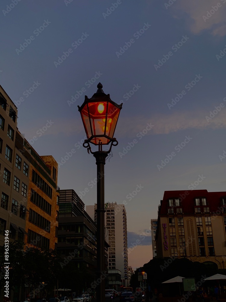 A glowing street lamp illuminates the twilight sky, framed by urban buildings on either side. The warm light contrasts with the fading daylight, creating a peaceful cityscape scene A glowing street lamp illuminates the twilight sky, framed by urban buildings on either side. The warm light contrasts with the fading daylight, creating a peaceful cityscape scene