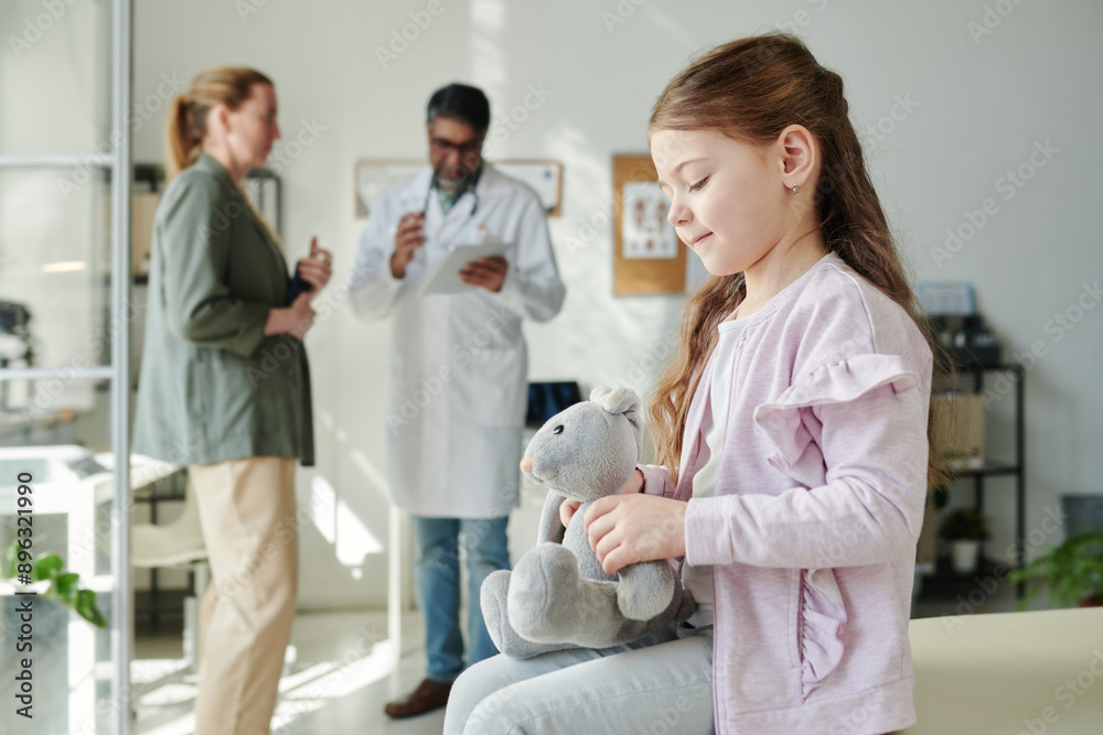 Fototapeta premium Side view of cute girl with grey soft toy sitting in front of camera in medical office and waiting for her mother consulting with clinician