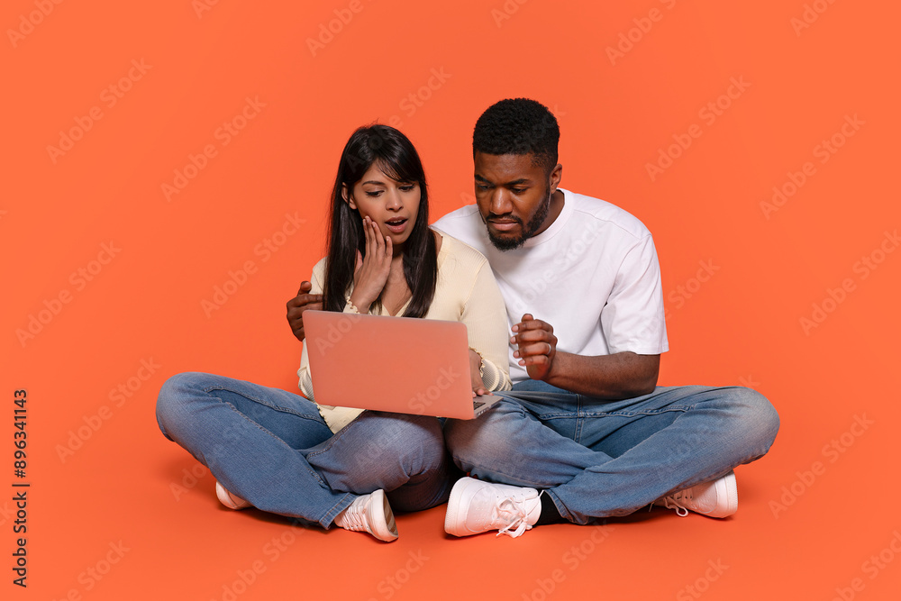 Shocked Couple Sitting on Orange Floor Looking at Laptop Shocked Couple Sitting on Orange Floor Looking at Laptop