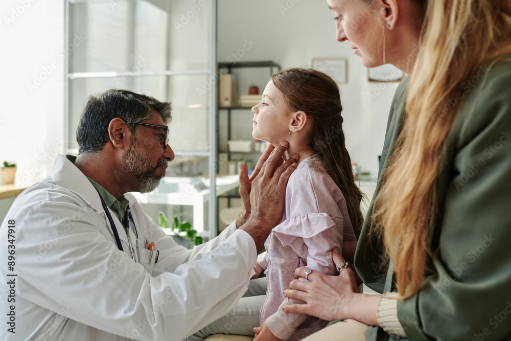 Side view of cute little patient of endocrinologist having her thyroid checked by experienced male clinician during consultation Side view of cute little patient of endocrinologist having her thyroid checked by experienced male clinician during consultation
