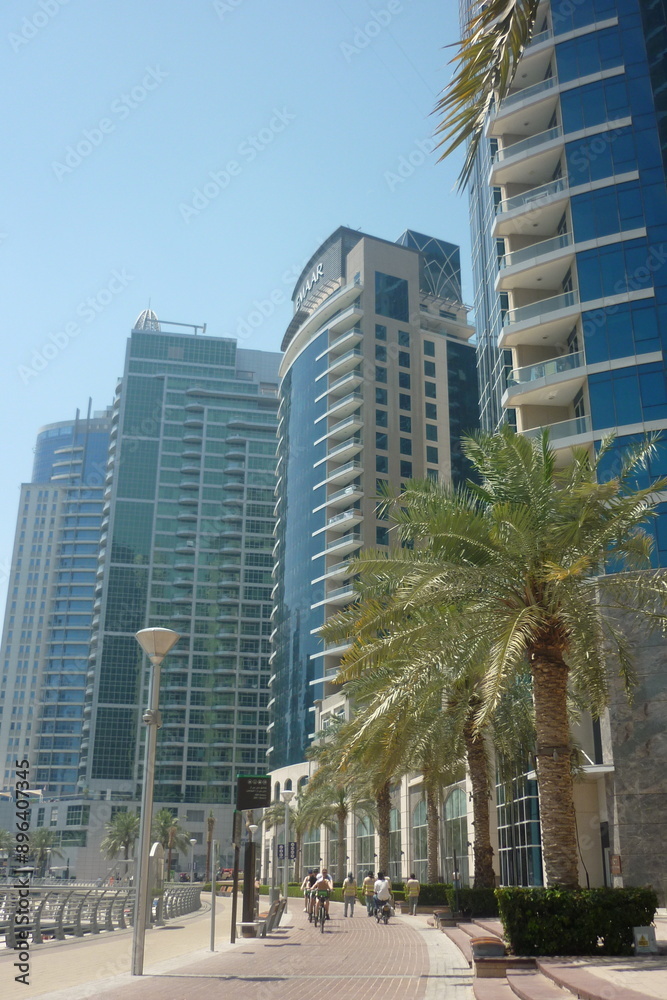 Modern skyscrapers with reflective glass facades line a sunny promenade in a bustling city. Palm trees, cyclists, and pedestrians create a vibrant, urban atmosphere. Modern skyscrapers with reflective glass facades line a sunny promenade in a bustling city. Palm trees, cyclists, and pedestrians create a vibrant, urban atmosphere.