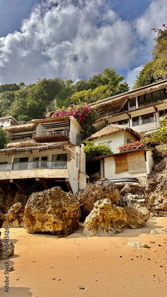 Fototapeta premium Charming houses on a rocky cliffside with thatched roofs, vibrant flowers, and lush greenery. Idyllic tropical setting with a sandy beach, perfect for serene coastal living and nature stock photos