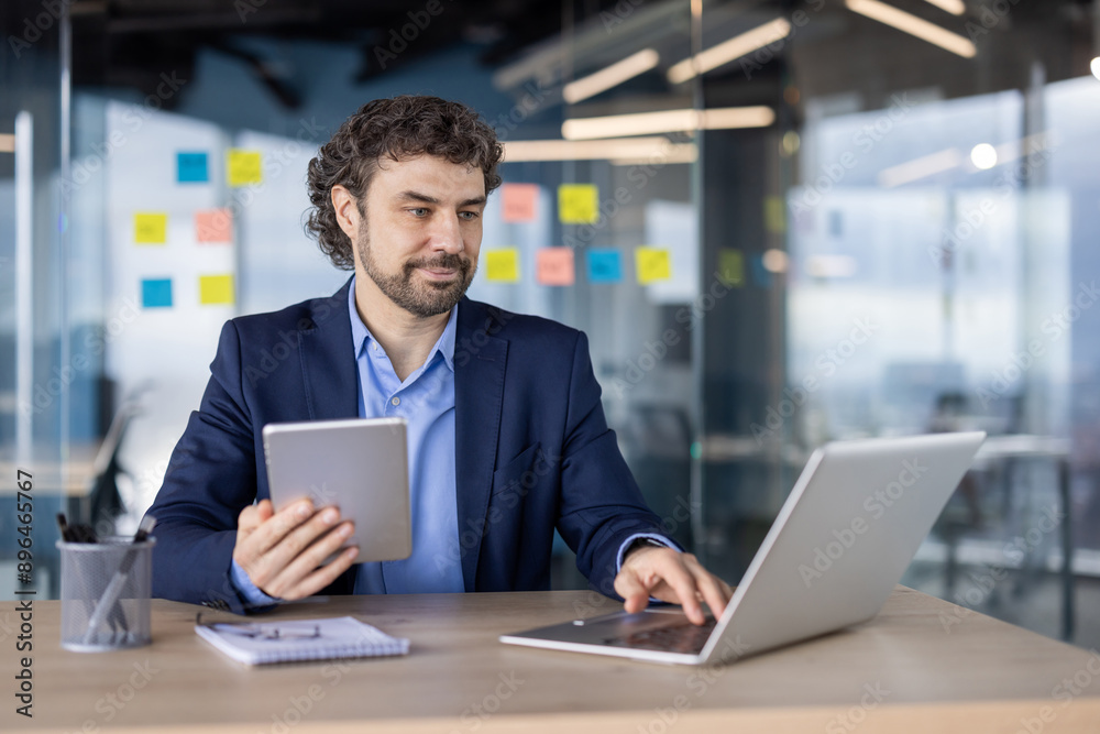 Businessman in formal attire working on laptop and holding tablet in modern office. Corporate workspace with sticky notes and glass walls. Professional environment with focused employee multitasking Businessman in formal attire working on laptop and holding tablet in modern office. Corporate workspace with sticky notes and glass walls. Professional environment with focused employee multitasking