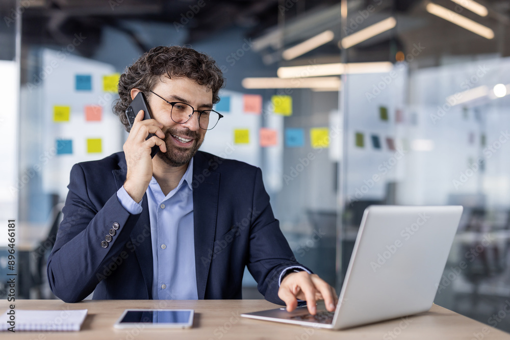 Businessman multitasking with laptop and phone in modern office. Professional in suit engaged in conversation and working on computer. Office environment with sticky notes in background. Businessman multitasking with laptop and phone in modern office. Professional in suit engaged in conversation and working on computer. Office environment with sticky notes in background.