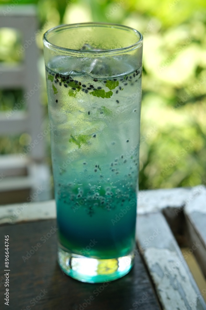A refreshing blue butterfly pea flower soda with basil seeds in a tall glass, placed on a wooden table with a lush green background. Perfect for summer refreshment, beverage advertisements, A refreshing blue butterfly pea flower soda with basil seeds in a tall glass, placed on a wooden table with a lush green background. Perfect for summer refreshment, beverage advertisements,
