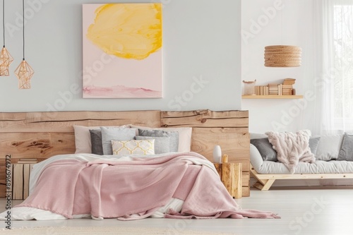 white and pink bedroom with wooden headboard, light grey floor, large pastel yellow abstract painting on the wall above bed frame, wood log stack side table, gray sofa in corner of room photo