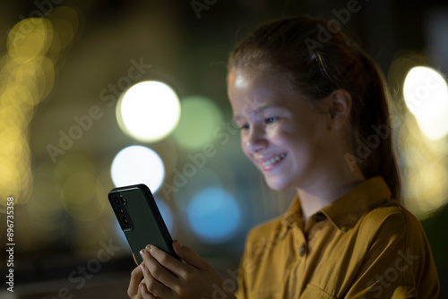 Positive child girl playing online game on her mobile phone with a friend outdoors at night