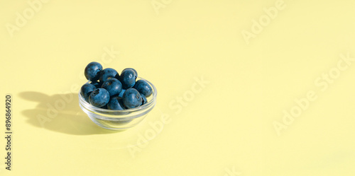 Fresh Blueberries in a glass bowl on a light yellow background, minimal summer fruits concept, banner with copy space