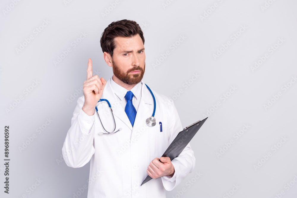 Portrait of experienced doctor man point finger hold clipboard wear white coat isolated on grey color background